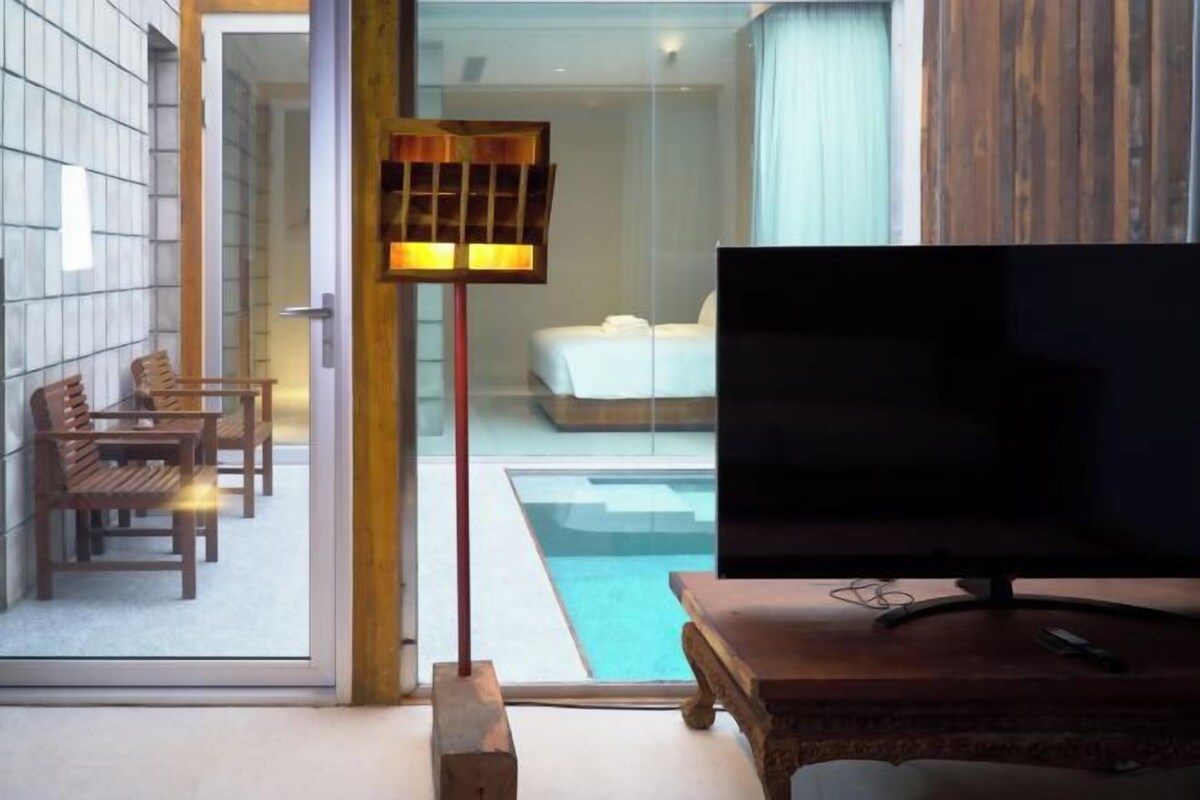 Quarto de hotel moderno com uma televisão grande, mobiliário de madeira, porta de vidro que dá para uma varanda com piscina e uma cama com roupa de cama branca.
