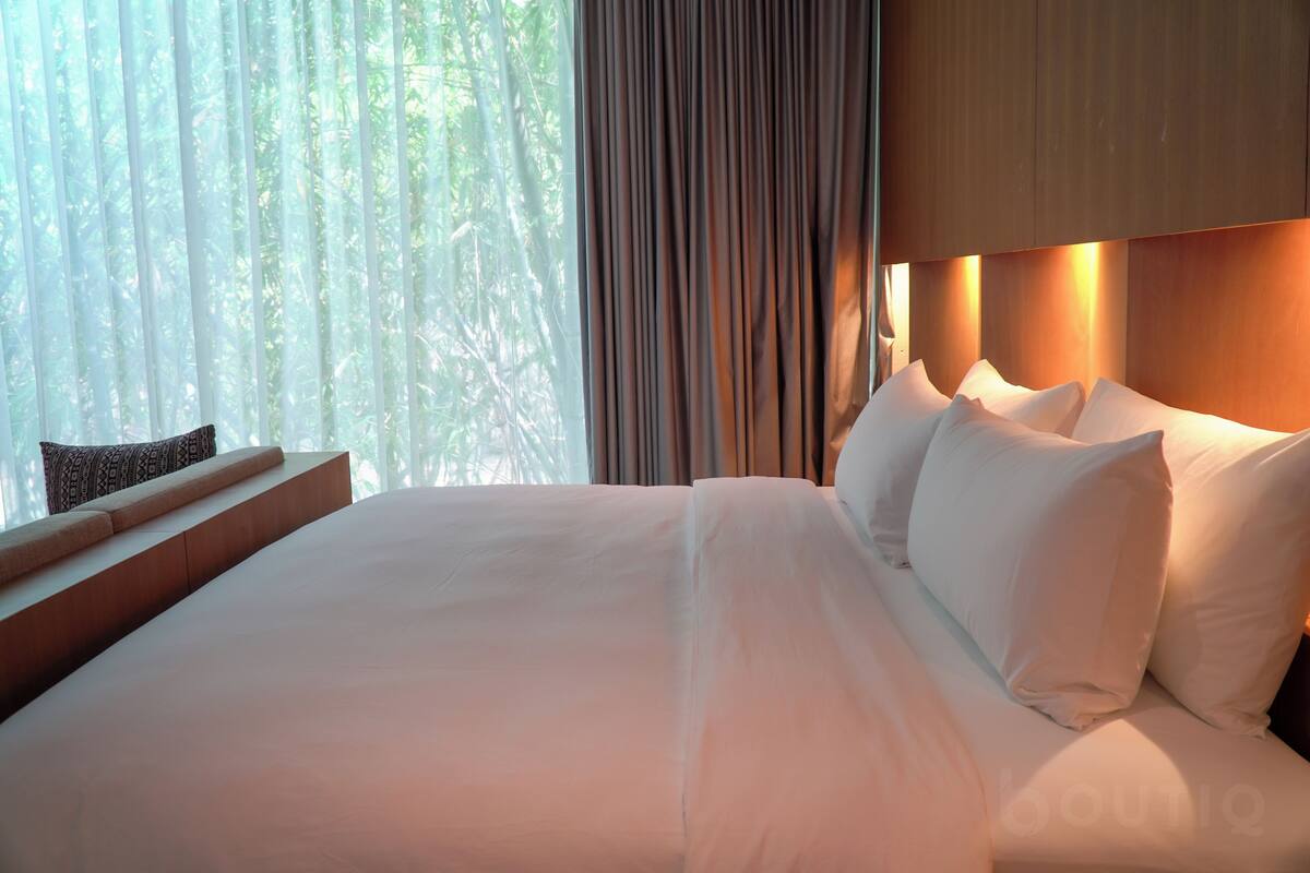 A neatly made bed with white linens and pillows, a wooden headboard, and a window with sheer curtains allowing natural light.