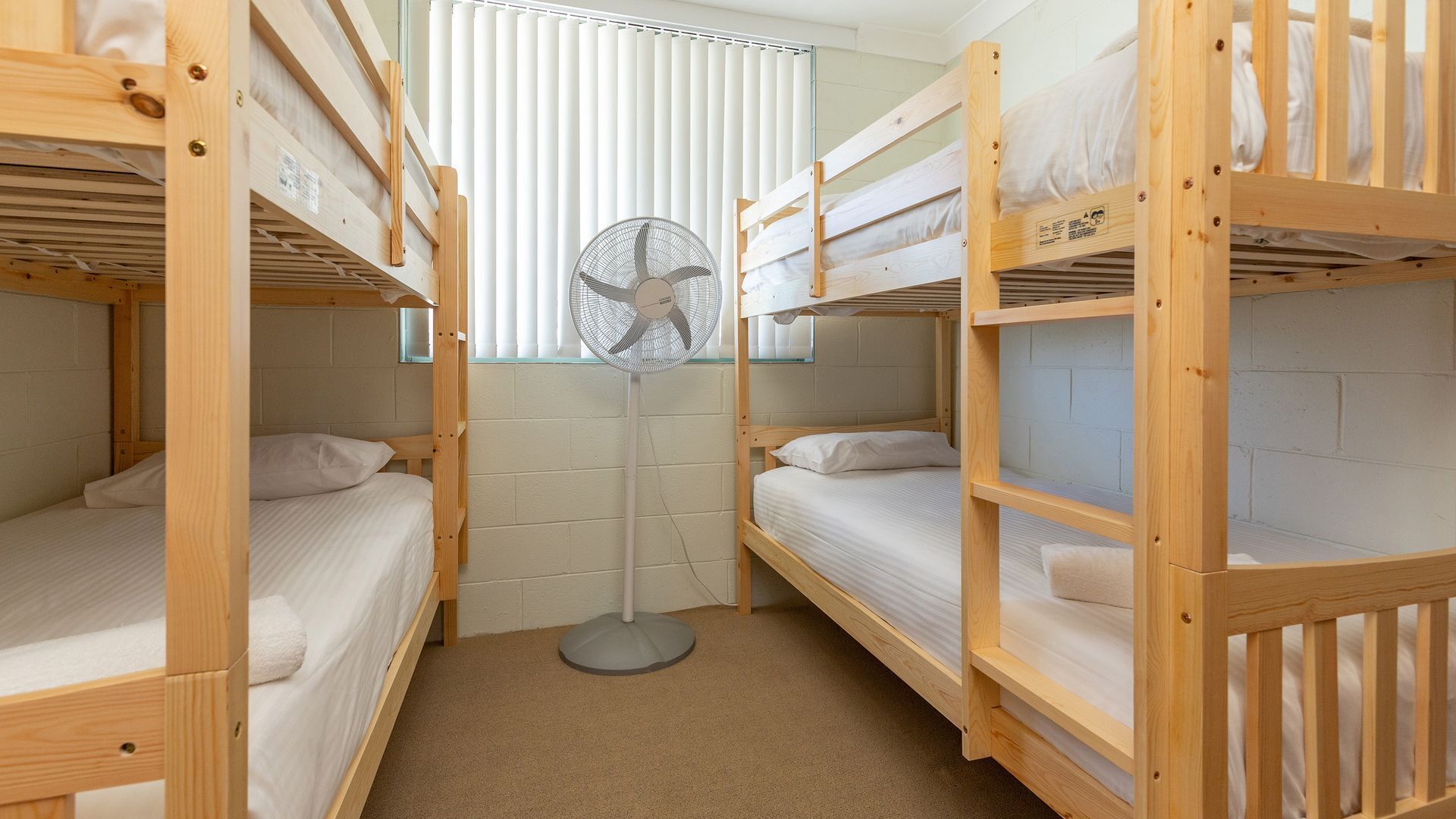 A dormitory room with bunk beds, a standing fan, and a window with blinds.