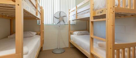 A dormitory room with bunk beds, a standing fan, and a window with blinds.