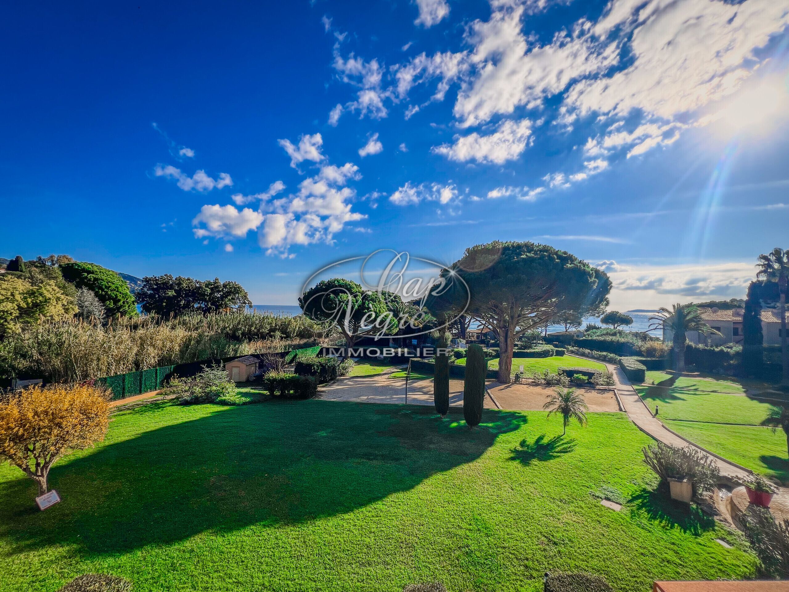 Um jardim bem cuidado com céu claro, uma árvore grande e uma placa para 'Negole Immobiliere'.