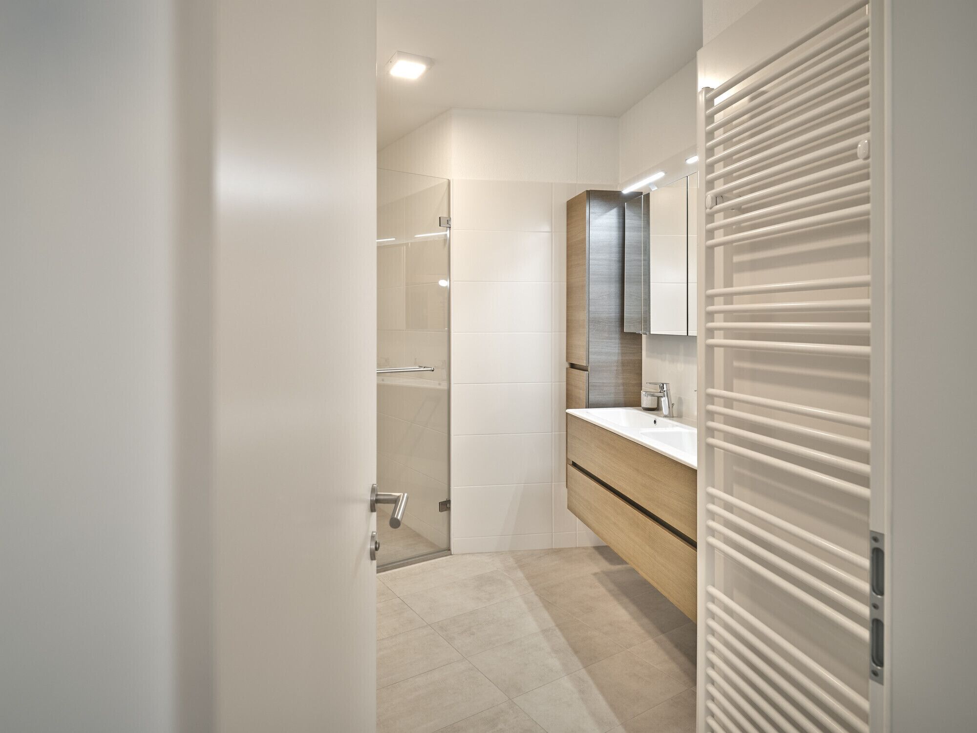 Un baño moderno con ducha de vidrio, tocador de madera y suelo de baldosas blancas.