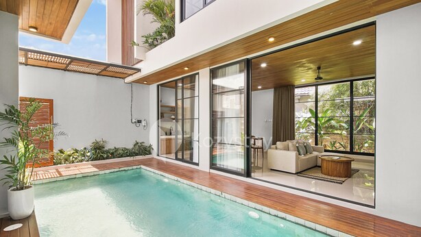 Un área de piscina moderna con cubierta de madera, un conjunto de sofás y grandes ventanales con vistas a la vegetación.