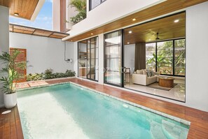 A modern pool area with a wooden deck, a sofa set, and large windows overlooking greenery.