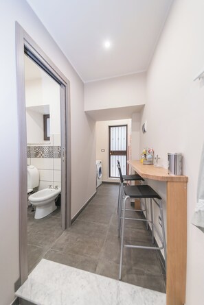 Un couloir étroit mène à une salle de bain équipée de toilettes et d’un bidet, ainsi que d’un lave-linge et d’un petit coin bar avec des tabourets.