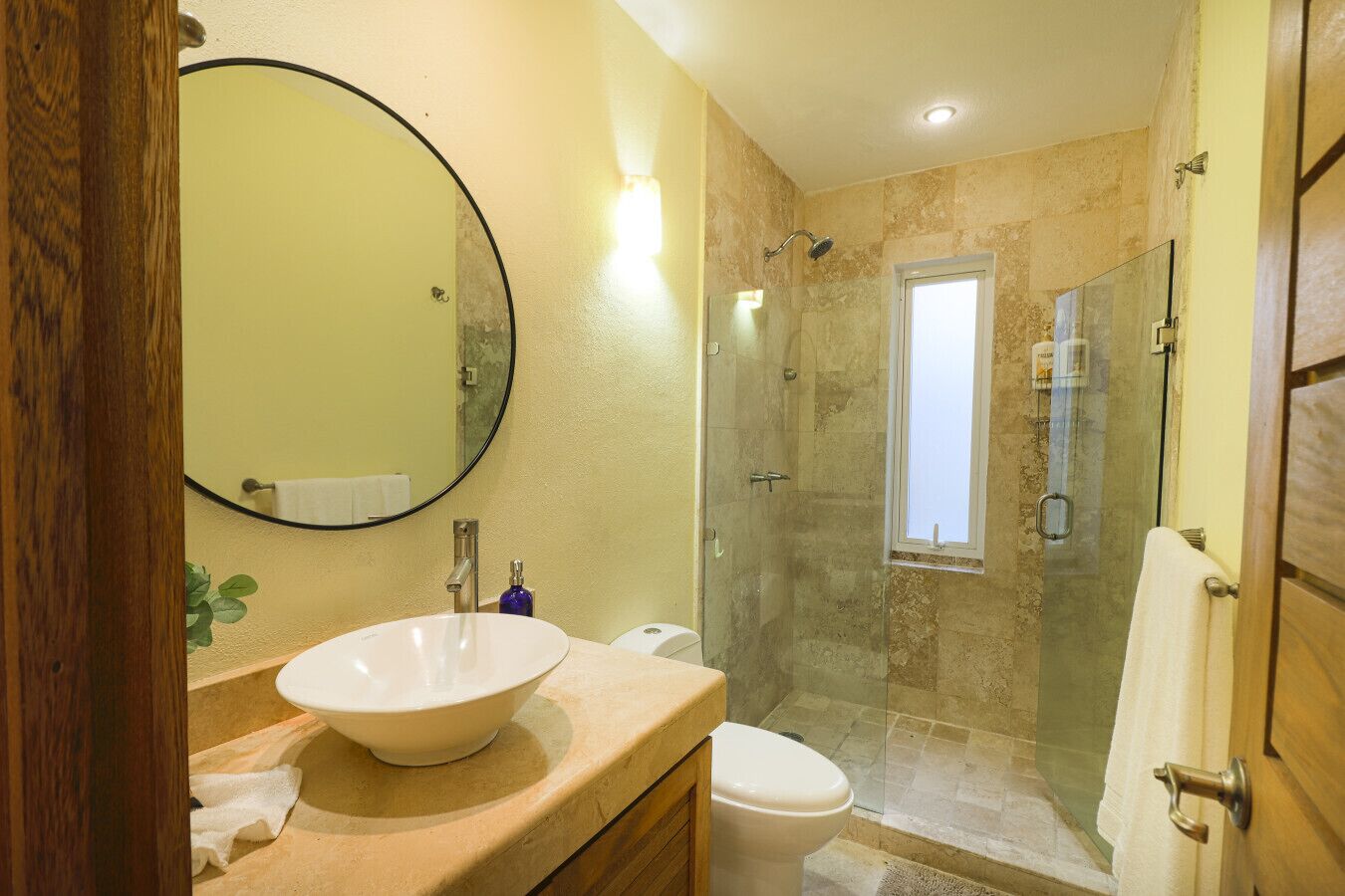 A bathroom with a large round mirror, a white sink, a toilet, and a glass-enclosed shower area.