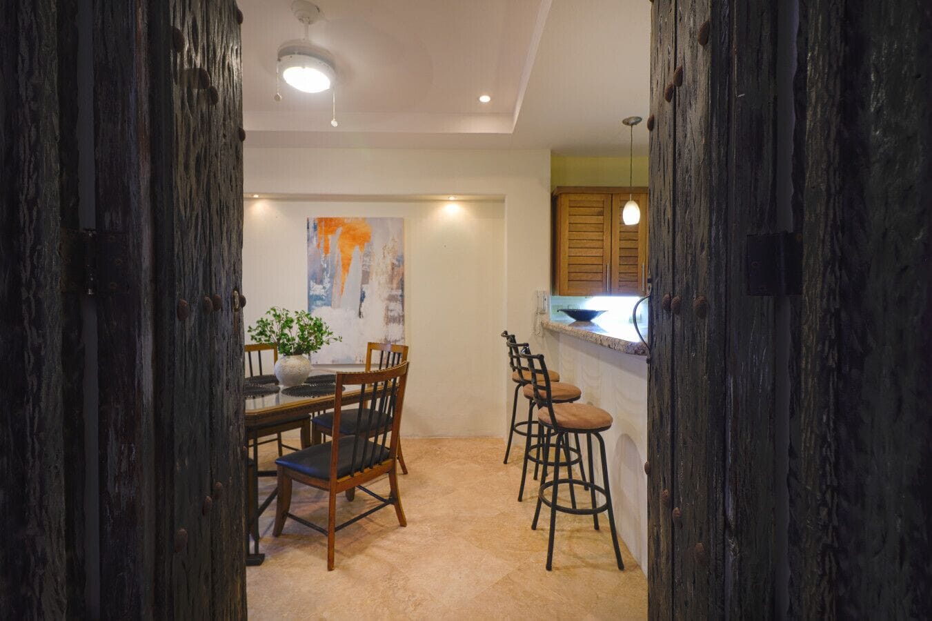 A dining area with a table and chairs, a painting on the wall, and a kitchen area with stools.