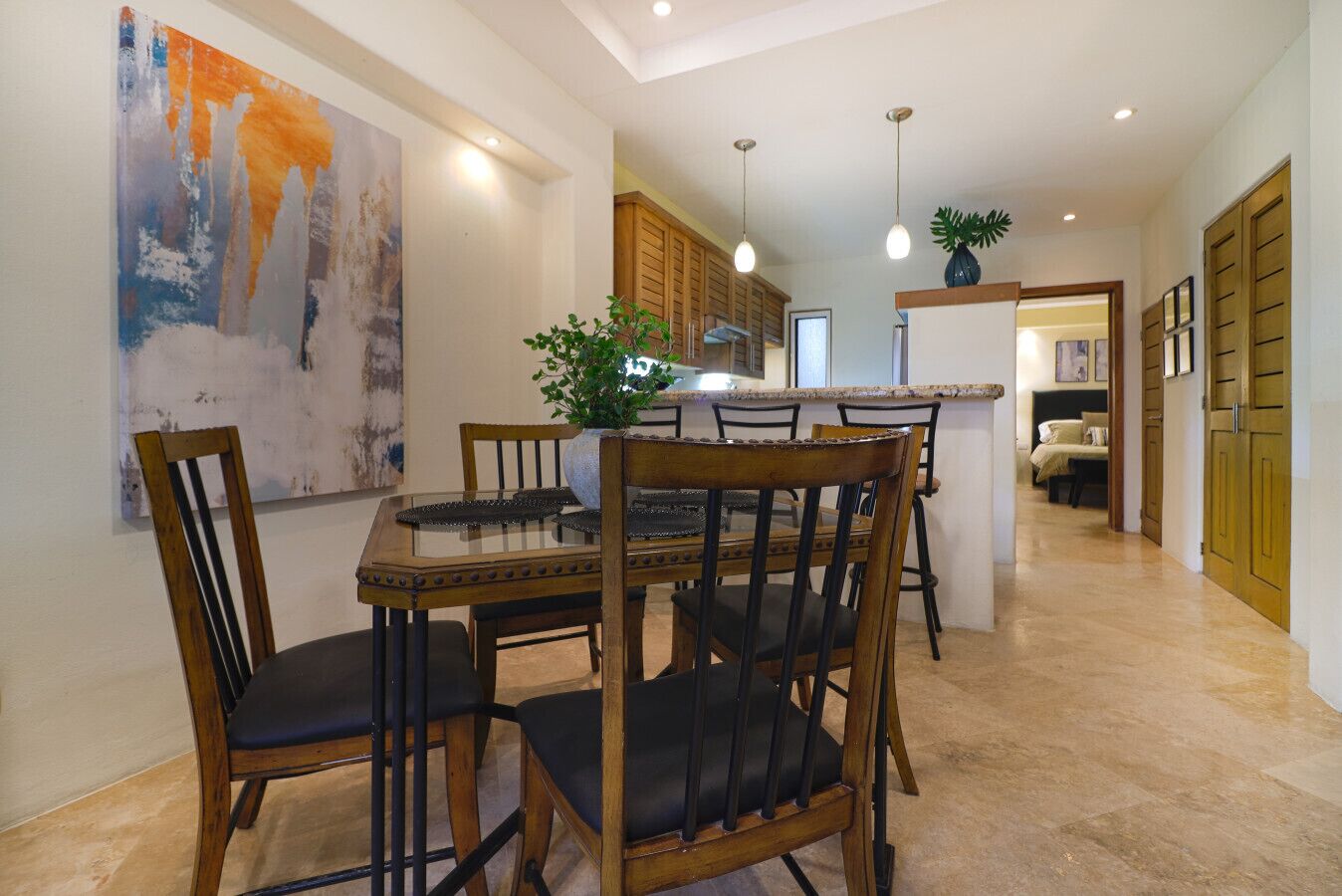 A modern kitchen with wooden cabinets, a dining table with chairs, pendant lights, and a large abstract painting on the wall.