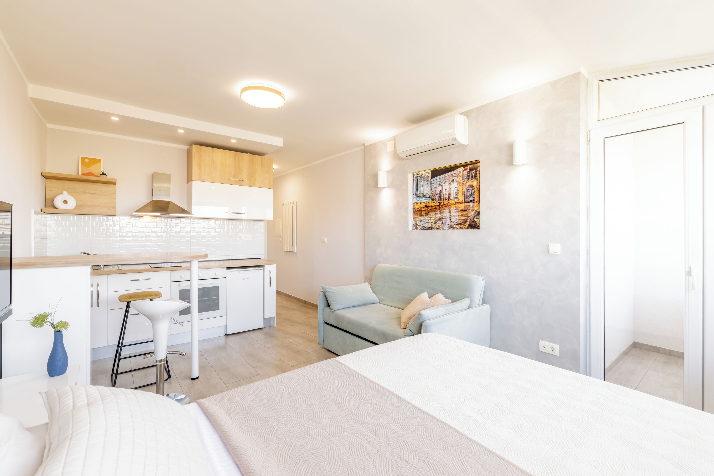 A modern hotel room with a kitchenette, a bed, a sofa, and a painting on the wall.