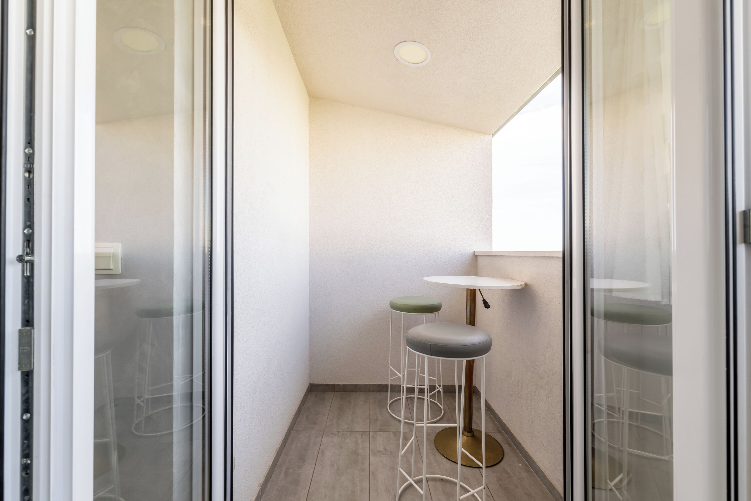 Een smalle, moderne badkamer met een glazen deur, een witte wastafel en twee krukjes.