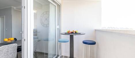 A modern balcony with a glass door leading to a dining area, a small round table with stools, and a cityscape view.
