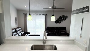 A modern kitchen with a stainless steel sink, white cabinets, a black leather sofa, and a ceiling fan.