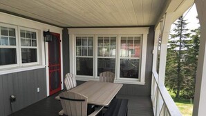 A covered porch with a wooden table and chairs, a red door, and large windows.