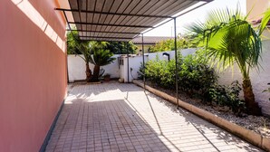A covered patio area with tiled flooring, a palm tree, and a wall with a white and yellow facade.