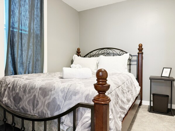 A neatly made bed with a patterned bedspread, a wooden headboard, and a bedside table with a picture frame and a trash bin.