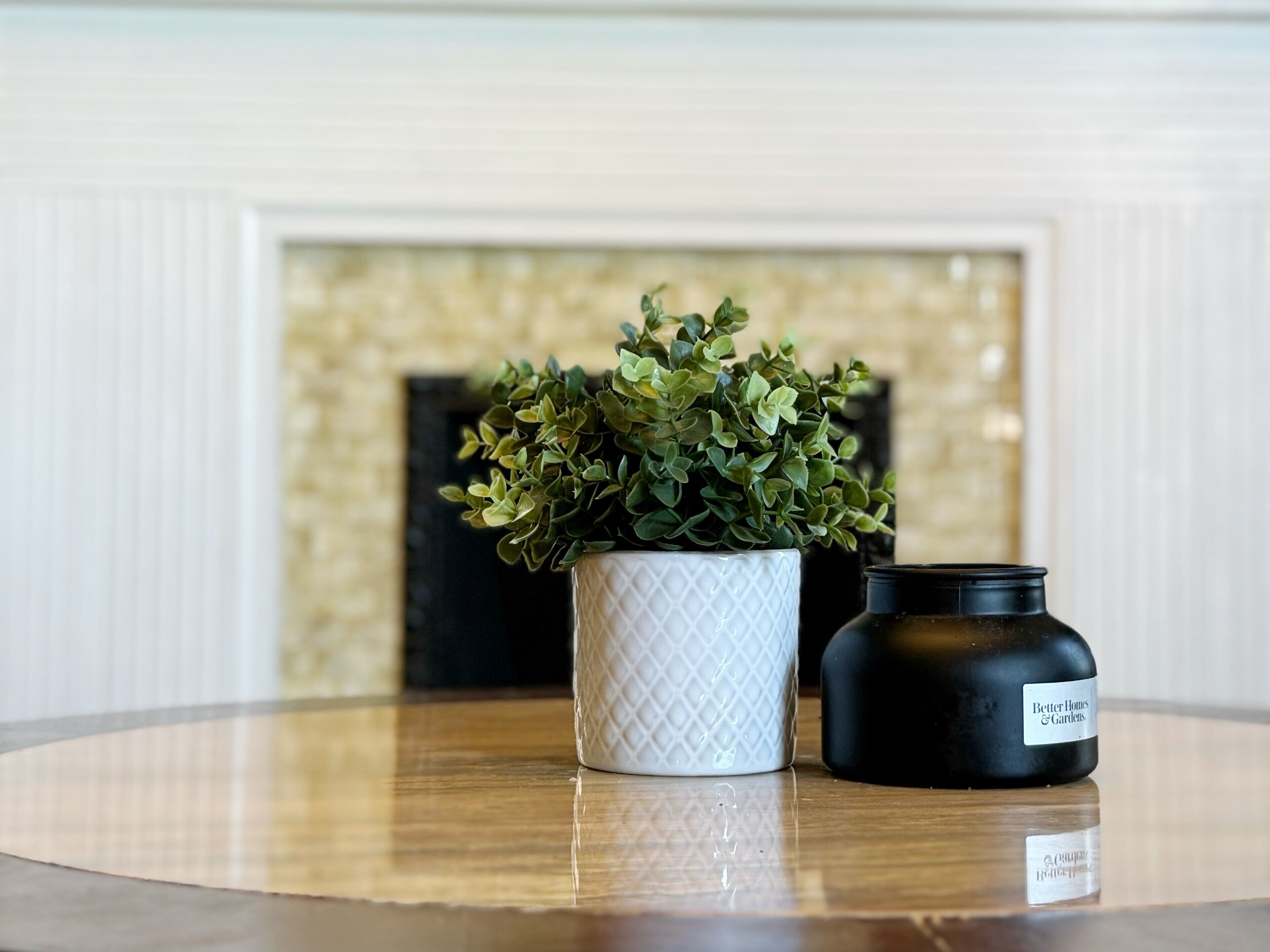 Una maceta blanca texturizada con una planta verde sobre una mesa de madera. Una vela negra con una etiqueta está al lado.