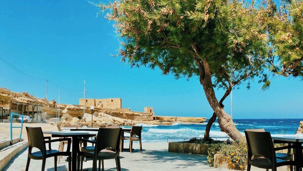 An outdoor seating area with tables and chairs, overlooking a coastal landscape with a view of the sea and a historic building in the background. - Chic Nadur Townhouse with Private Pool & BBQ (In-Nadur)
