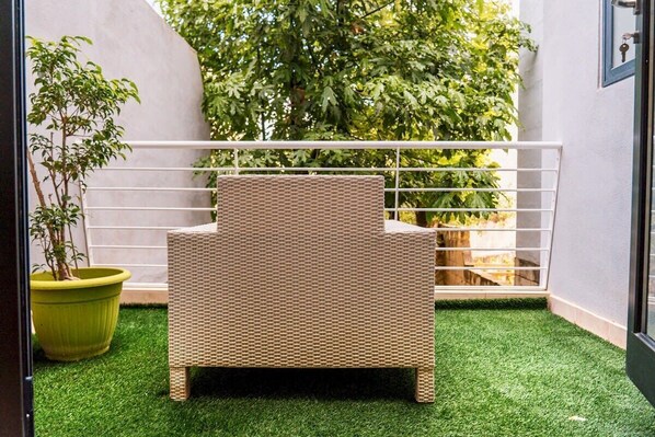 Una silla de mimbre en un espacio exterior pequeño y cerrado, con césped artificial, una planta en maceta y una barandilla blanca.