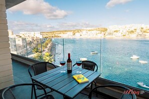 A balcony with a table, two chairs, a bottle of wine, and a glass of red wine. A book titled "Anything is Possible" is on the table. The view includes a body of water with boats and a coastal town in the background.