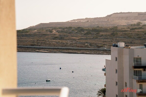 A coastal view with a building overlooking a calm sea, a rocky hillside, and a clear sky.