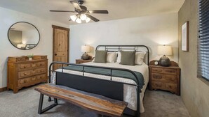 Une chambre à coucher comprenant un lit, une commode en bois, des tables de chevet, un banc, un ventilateur de plafond et un miroir.