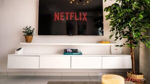 Una sala moderna con un televisor de pantalla plana montado en una pared blanca, una planta en maceta y un estante con libros y una caja de juegos.