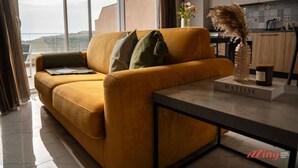 Un salon moderne avec un canapé de couleur moutarde, une table basse et un vase contenant des plumes d’herbe des pampas. Une bouteille de vin et un livre intitulé « MATISSE » sont posés sur la table. La pièce est dotée de grandes fenêtres et offre une vue sur l’extérieur.