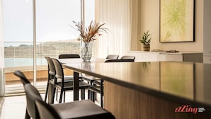Un espace de repas moderne avec une longue table, des chaises et un vase rempli de plantes séchées. Une grande fenêtre offre une vue sur l’extérieur.