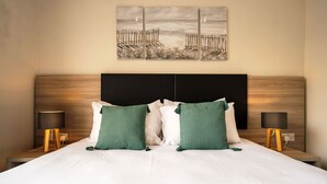 Un lit bien fait, avec du linge de lit blanc et des coussins verts, est encadré de deux tables de chevet sur lesquelles sont posées des lampes. Une grande œuvre d’art représentant une jetée et un voilier est accrochée au-dessus du lit.