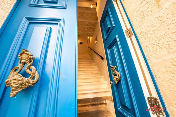 Una puerta azul con elegantes detalles dorados, que da acceso a una escalera con escalones blancos y una pared color crema.