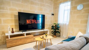 Una sala de estar con una televisión de pantalla plana grande mostrando 'NETFLIX', una mesa de centro de madera, una planta en maceta y un sofá.