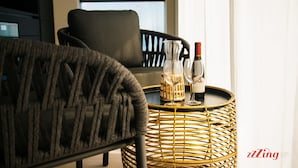 Una habitación con una silla de mimbre, una mesa redonda con una botella y vasos, y una ventana con vistas.