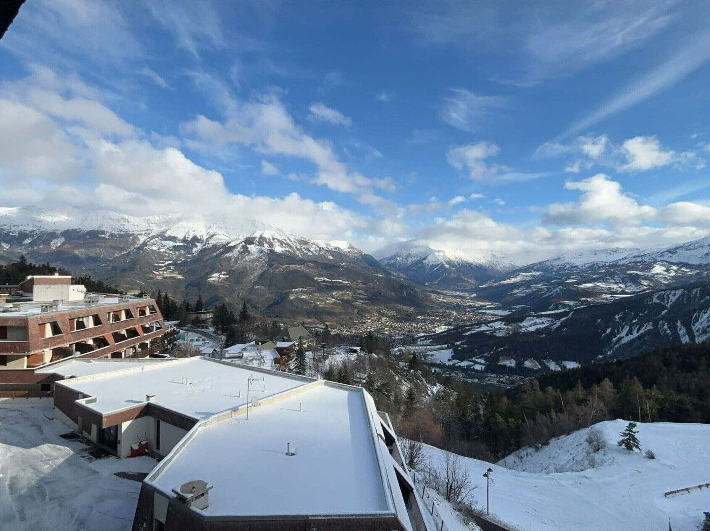 A snow-covered mountain landscape with a town in the valley and a building with a flat roof in the foreground.