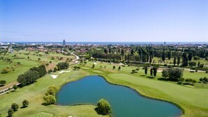 Campo de golf con un estanque, árboles y un horizonte urbano distante.