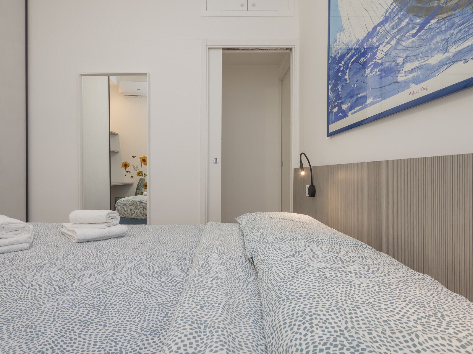 A bedroom with a bed, a nightstand, a wall-mounted lamp, a painting, and a door leading to a bathroom.