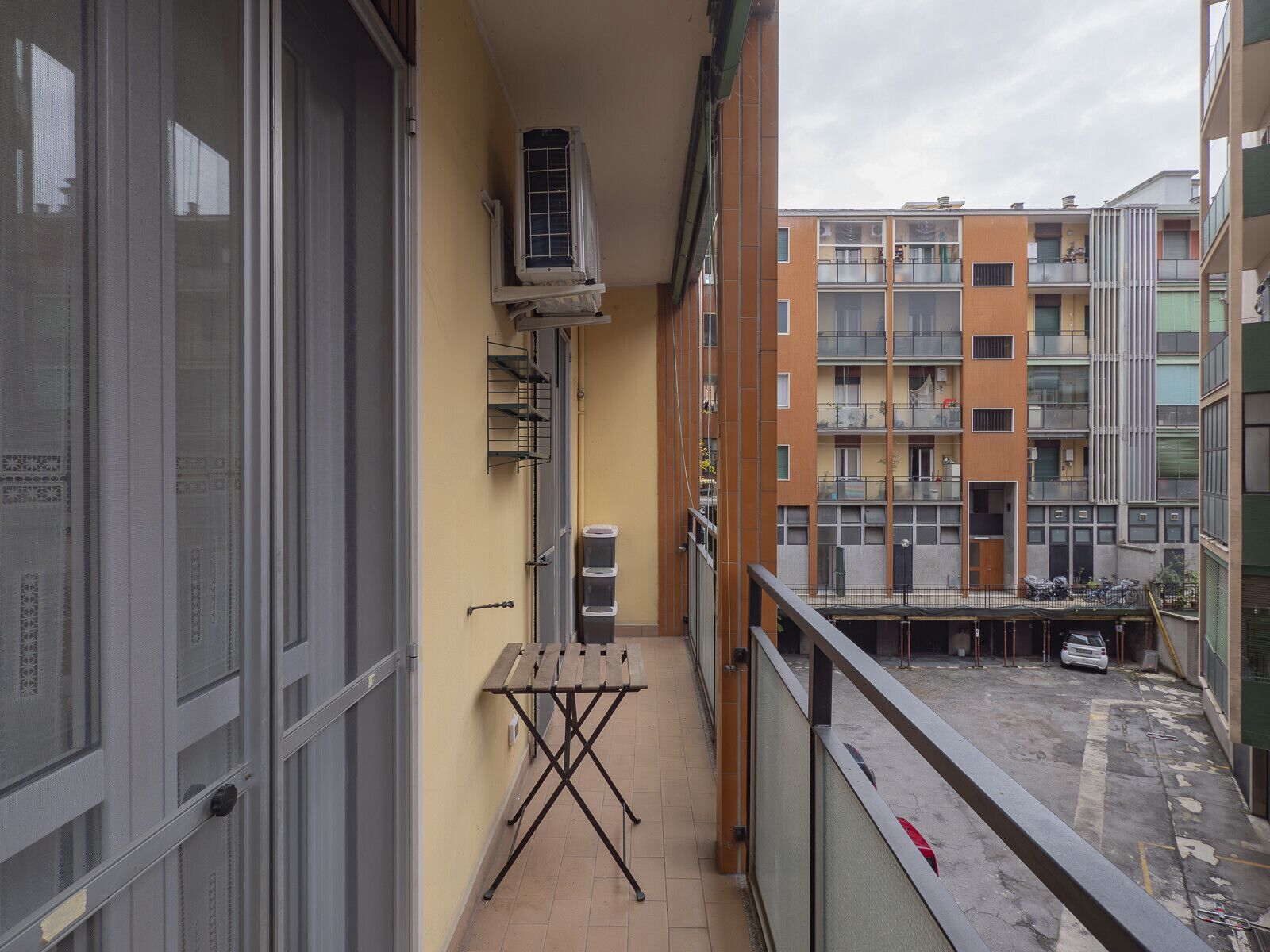 Un balcón con mesa de madera, unidad de aire acondicionado y vista a edificios de varios pisos.