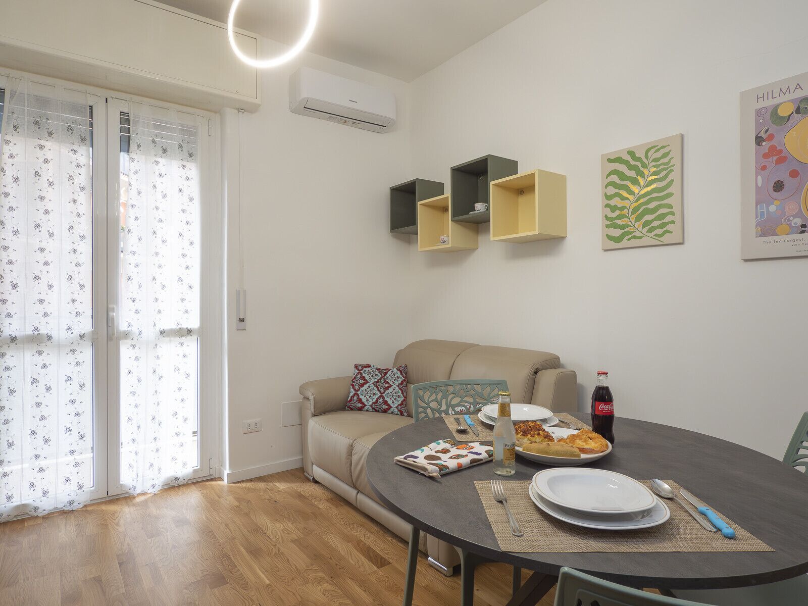 A modern living room with a beige sofa, a dining table with plates and cutlery, and a small kitchen area with cabinets.