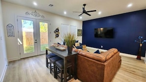A modern living room with a brown leather sofa, a dining table with chairs, a ceiling fan, and a TV mounted on the wall.