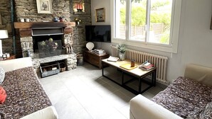 A cozy living room with a stone fireplace, a television, and a large window with a view of greenery. - Family house near Groix beach, garden and garage (Groix)