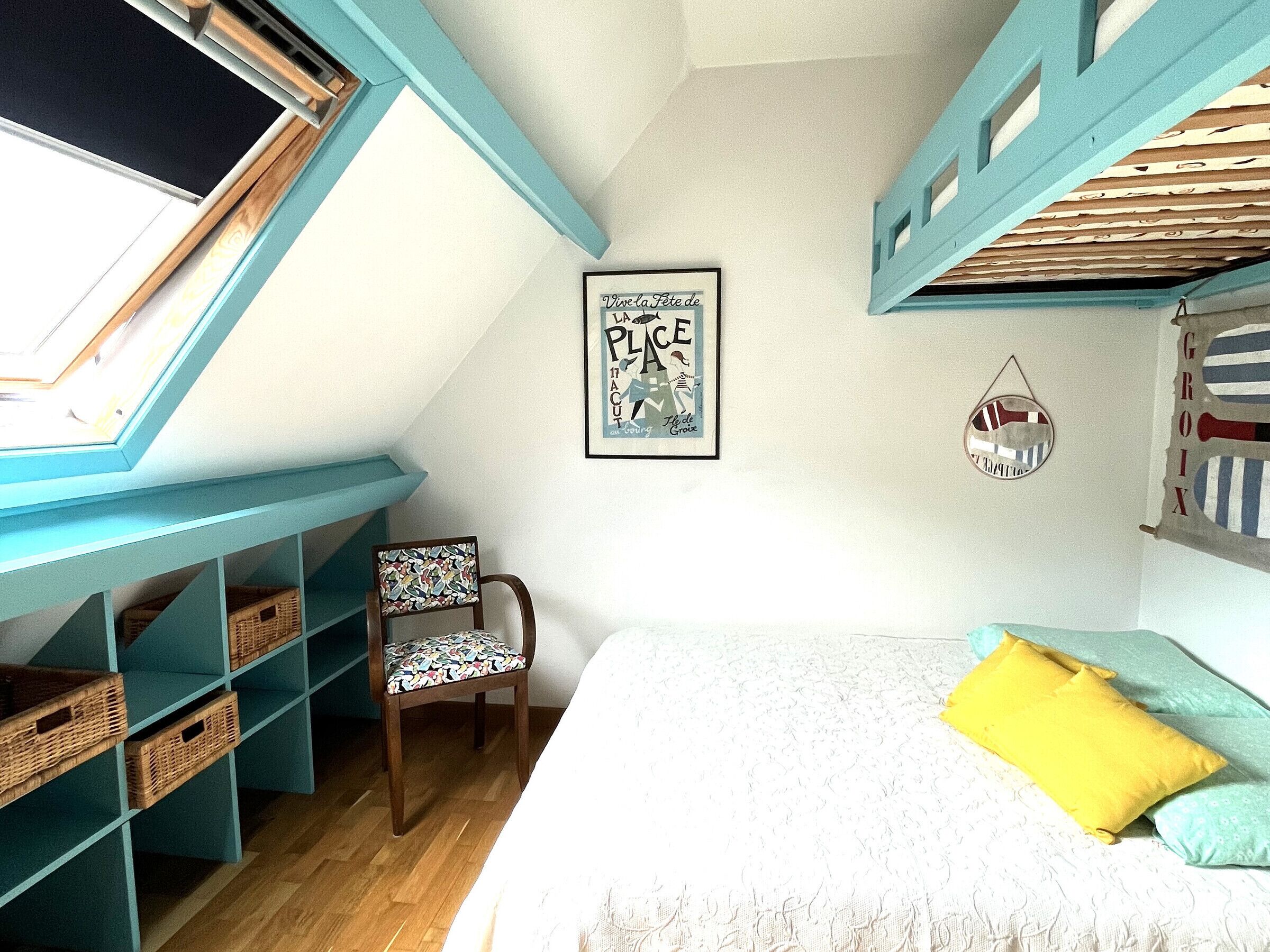 A bedroom with a bunk bed, a chair, a bed with a yellow pillow, a wall with a framed poster, and a window with a skylight.