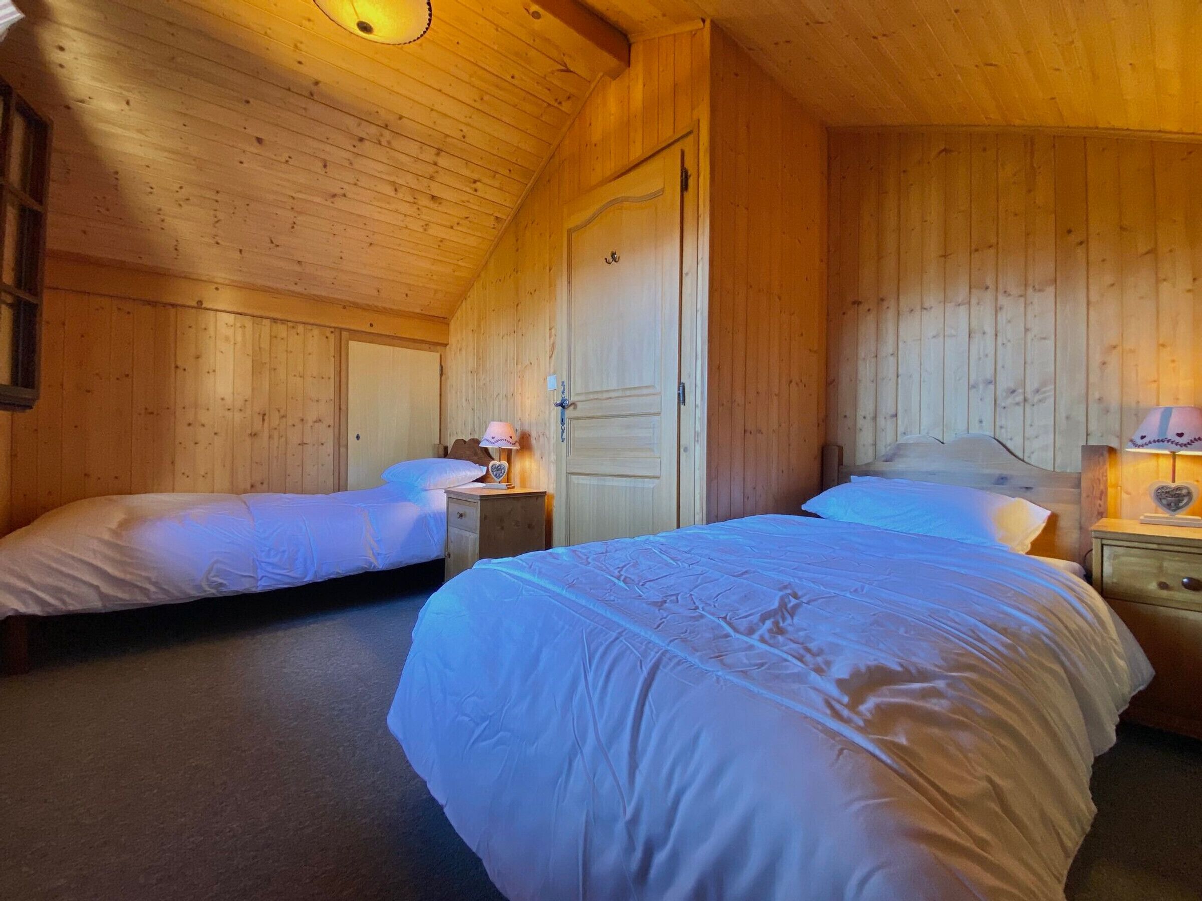 A cozy wooden room with two beds, a nightstand with a lamp, and a door.