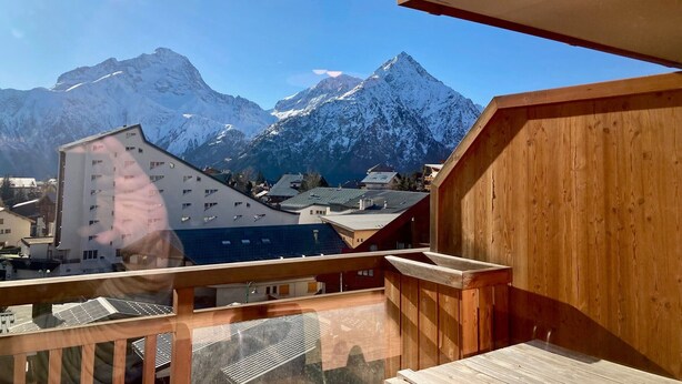 A wooden balcony with a view of snow-capped mountains and a building with multiple windows.