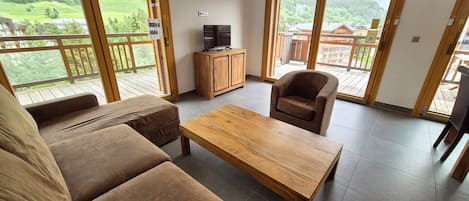 A spacious living area with a sofa, a wooden coffee table, and a chair. There is a television on a wooden cabinet, and large windows with a view of a green landscape.