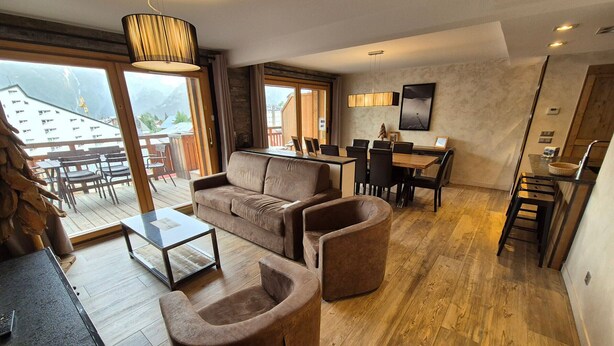 Ein modernes Wohnzimmer mit Sofa, Sesseln und Couchtisch. Es gibt einen Essbereich mit Tisch und Stühlen sowie eine Küchenzeile mit einer Bar. Große Fenster bieten Blick ins Freie.