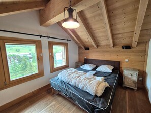 A bedroom with a wooden ceiling, a bed with white bedding, two windows with views of greenery, and a small bedside table.