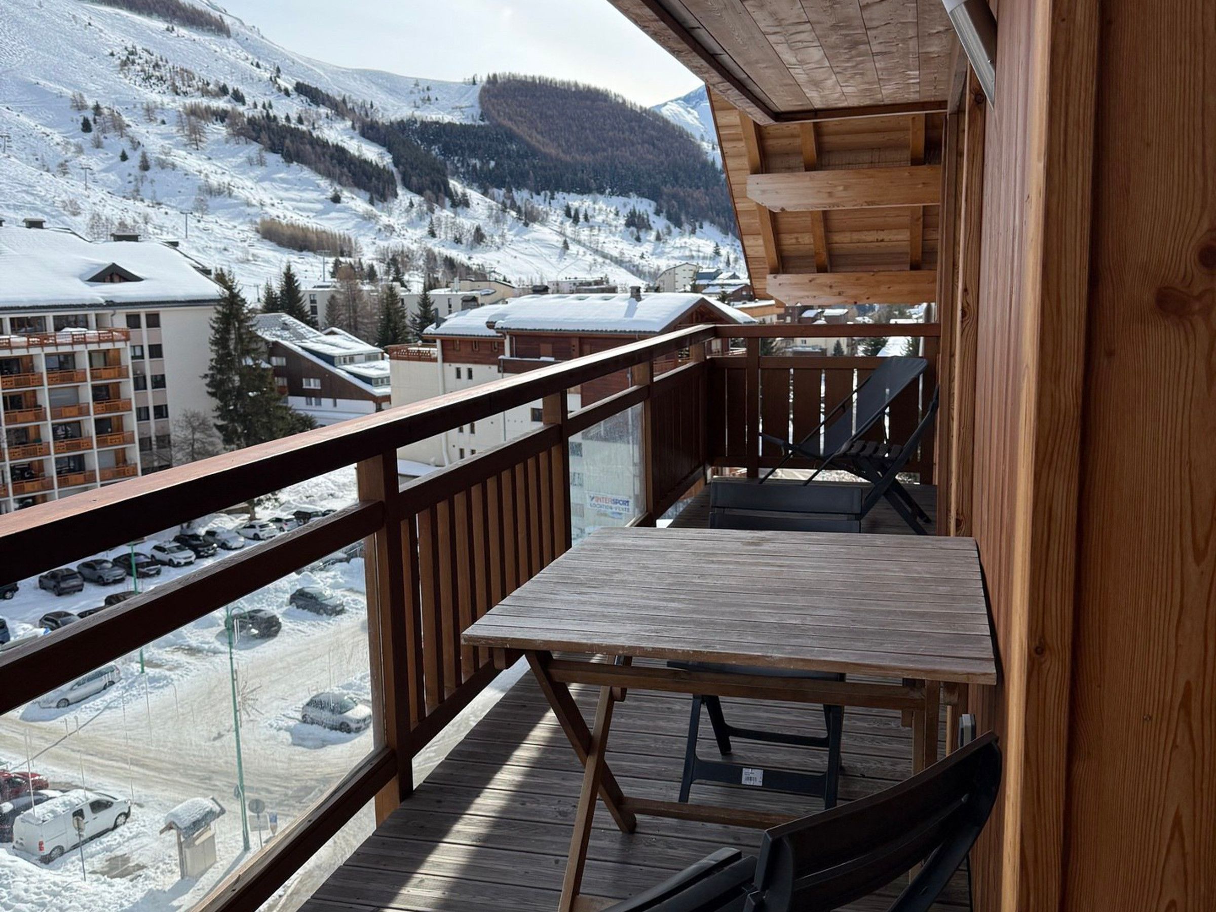 A wooden balcony with a table and chairs, overlooking a snowy mountain landscape and a parking area.