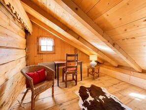 Ein gemütliches Dachzimmer mit Holzwänden, einem Fenster, einem Sessel, einem Tisch, einer Lampe und einem Kuhfellteppich.