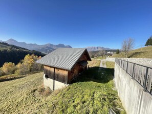 A wooden cabin with a sloped roof, situated on a grassy hillside with a stone wall and a path leading to it.