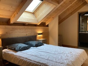 A bedroom with a skylight, wooden ceiling beams, a bed with white bedding and two pillows, a nightstand with a lamp, and a dresser. - Spacious bright apartment, 10 pers, pets allowed, 250m from slopes (Les Deux Alpes)