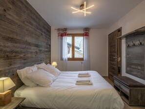 Ein Schlafzimmer mit einem hölzernen Kopfende, einem Bett mit weißer Bettwäsche, einem Fenster mit Vorhängen, einer Nachttischlampe und einem Holzschrank.